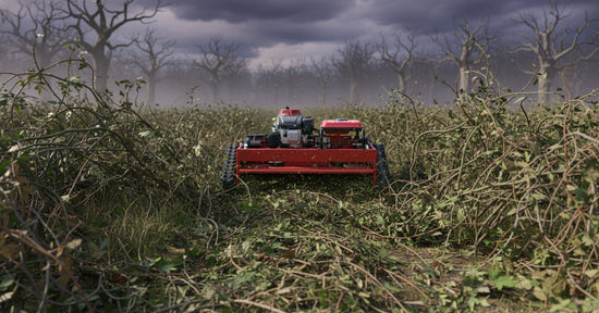 Red remote-controlled crawler lawn mower clearing dense, overgrown brush in a field under a dark, cloudy sky with bare trees 