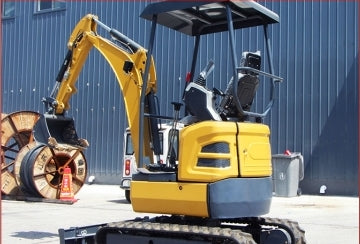 Side view of the JDSCT-25 compact excavator, yellow and black, with its arm extended, parked outside a building.